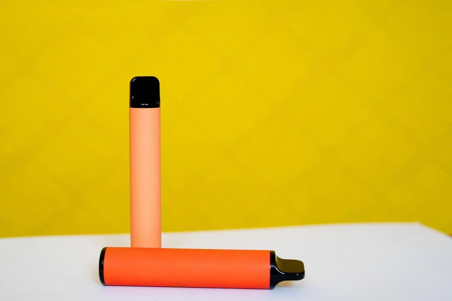 Two orange disposable pod-style vape devices on a white surface with yellow background