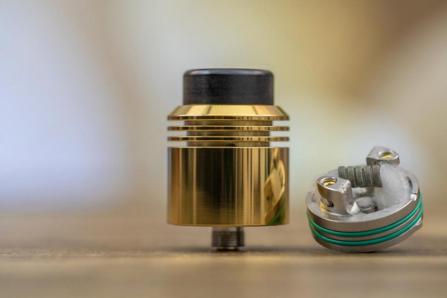 Golden rebuildable dripping atomizer next to a build deck with mesh coil, showing modern vape hardware